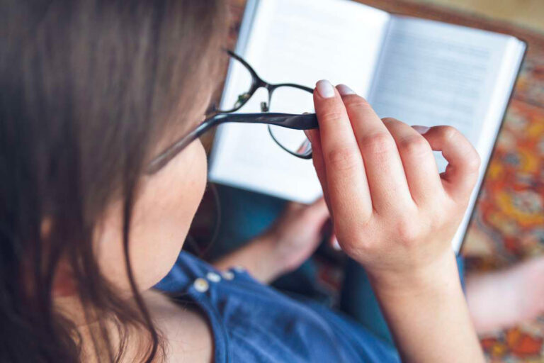 Dame brune assise dans son fauteuil en train de lire un livre avec ses lunettes de lecture